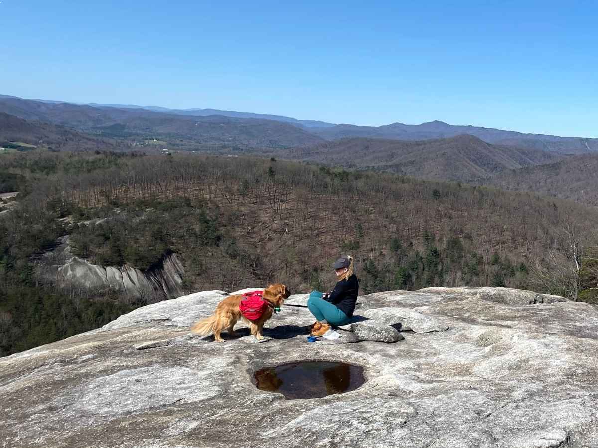 A woman and her golden retriever, who’s wearing a red dog backpack, rest on a rocky summit overlooking a vast mountain range under a clear blue sky. The scene reflects the reward and companionship found in backpacking with dogs through rugged terrain.