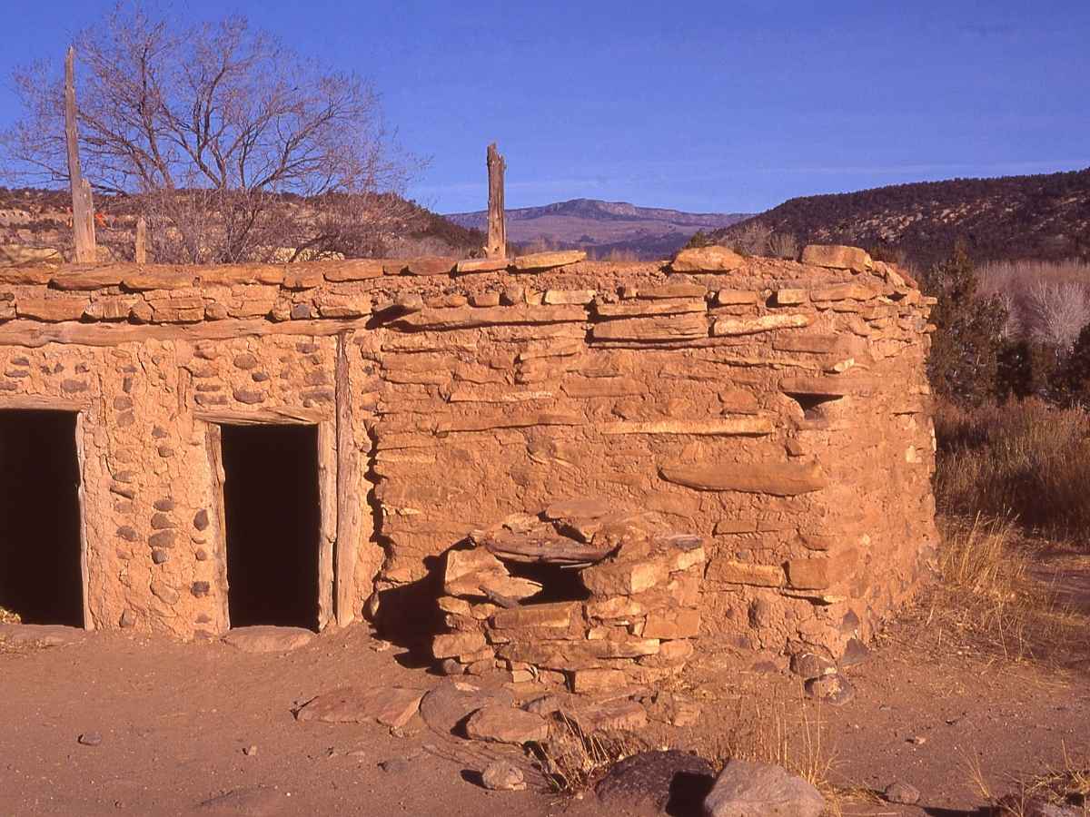 Small stone structure made of stacked sandstone and timber beams with two dark doorway openings in a dry desert landscape, a scenic stop on a Las Vegas to Zion National Park road trip. The historic Anasazi adobe building sits among scrub brush and rolling hills under a clear blue sky, showing traditional Southwestern architecture and rustic construction details.