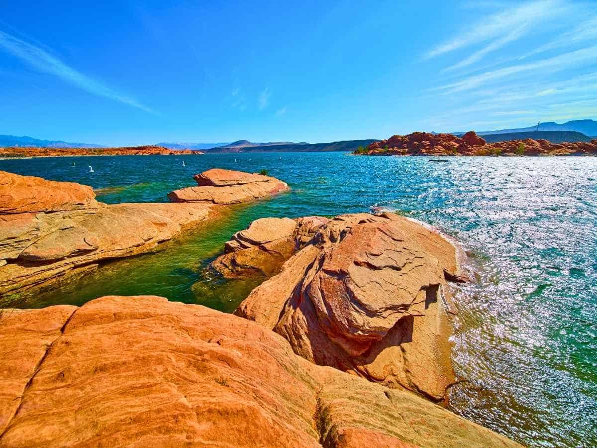 Bright blue lake at Sand Hollow State Park surrounded by smooth red sandstone rocks and desert hills, a scenic stop on a las vegas to zion national park road trip. The clear water sparkles under a sunny sky, contrasting with the warm orange rock formations along the shoreline.