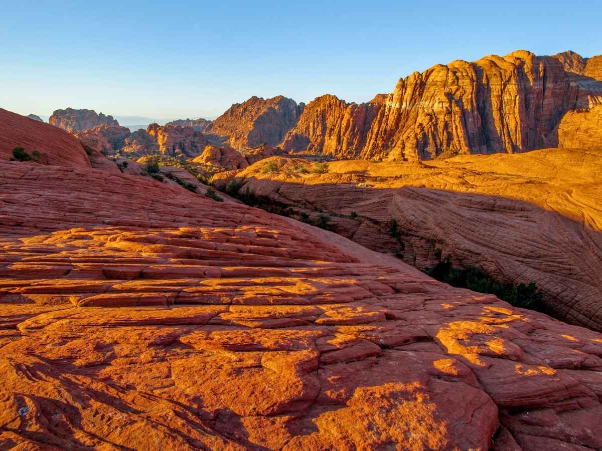 Expansive red and orange sandstone landscape glowing in golden light, a breathtaking highlight of a las vegas to zion national park road trip. Layered rock formations and towering cliffs stretch across the desert horizon under a clear blue sky, showcasing the dramatic scenery of the American Southwest.