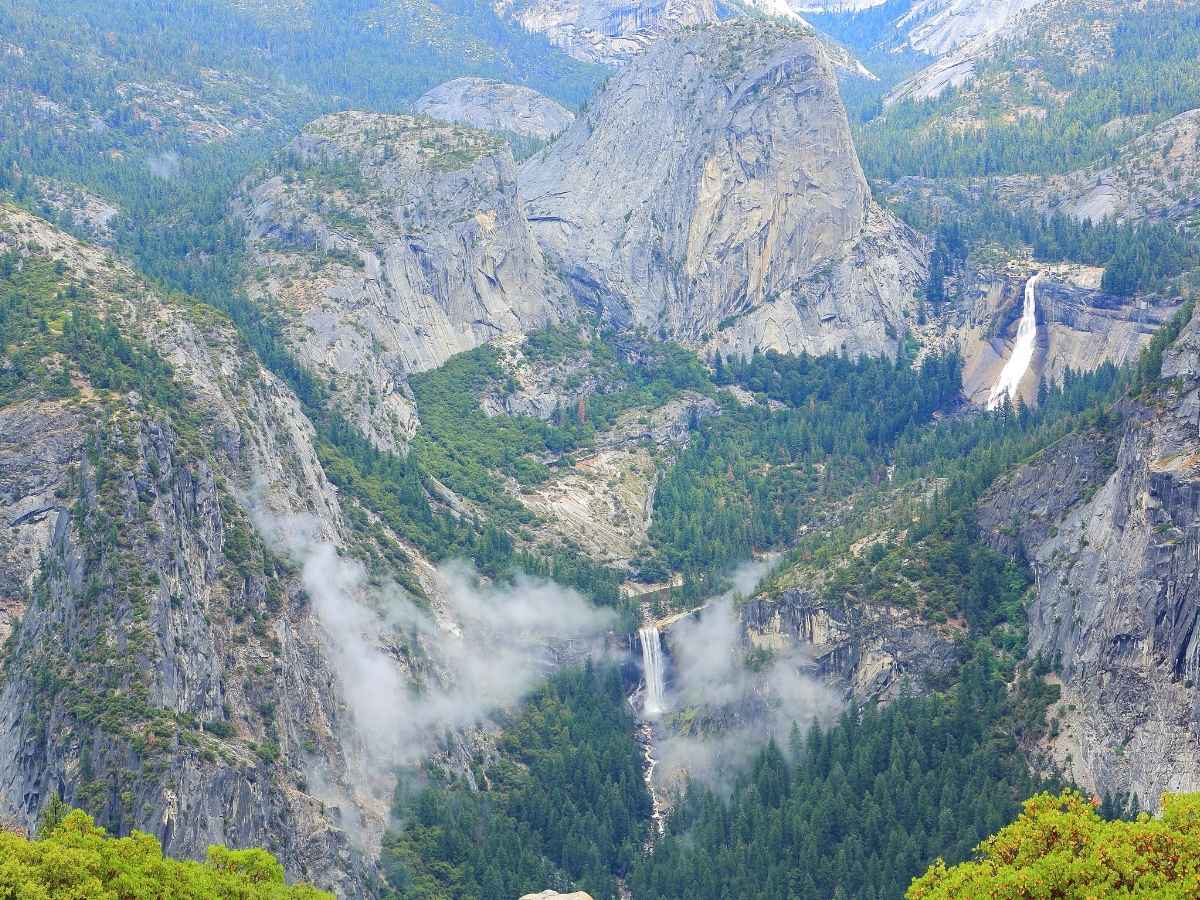 A sweeping aerial view of Yosemite Valley shows towering granite cliffs, dense pine forests, and multiple waterfalls cascading down into the canyon below. Low clouds drift through the valley, highlighting the dramatic and layered scenery to include in a Yosemite itinerary.