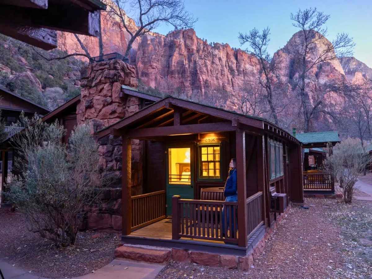 Cozy cabin with a lit porch and stone chimney set beneath towering red cliffs at dusk, a memorable stay on a las vegas to zion national park road trip. Warm light glows from the windows while rugged canyon walls rise dramatically in the background, highlighting scenic lodging near Zion.