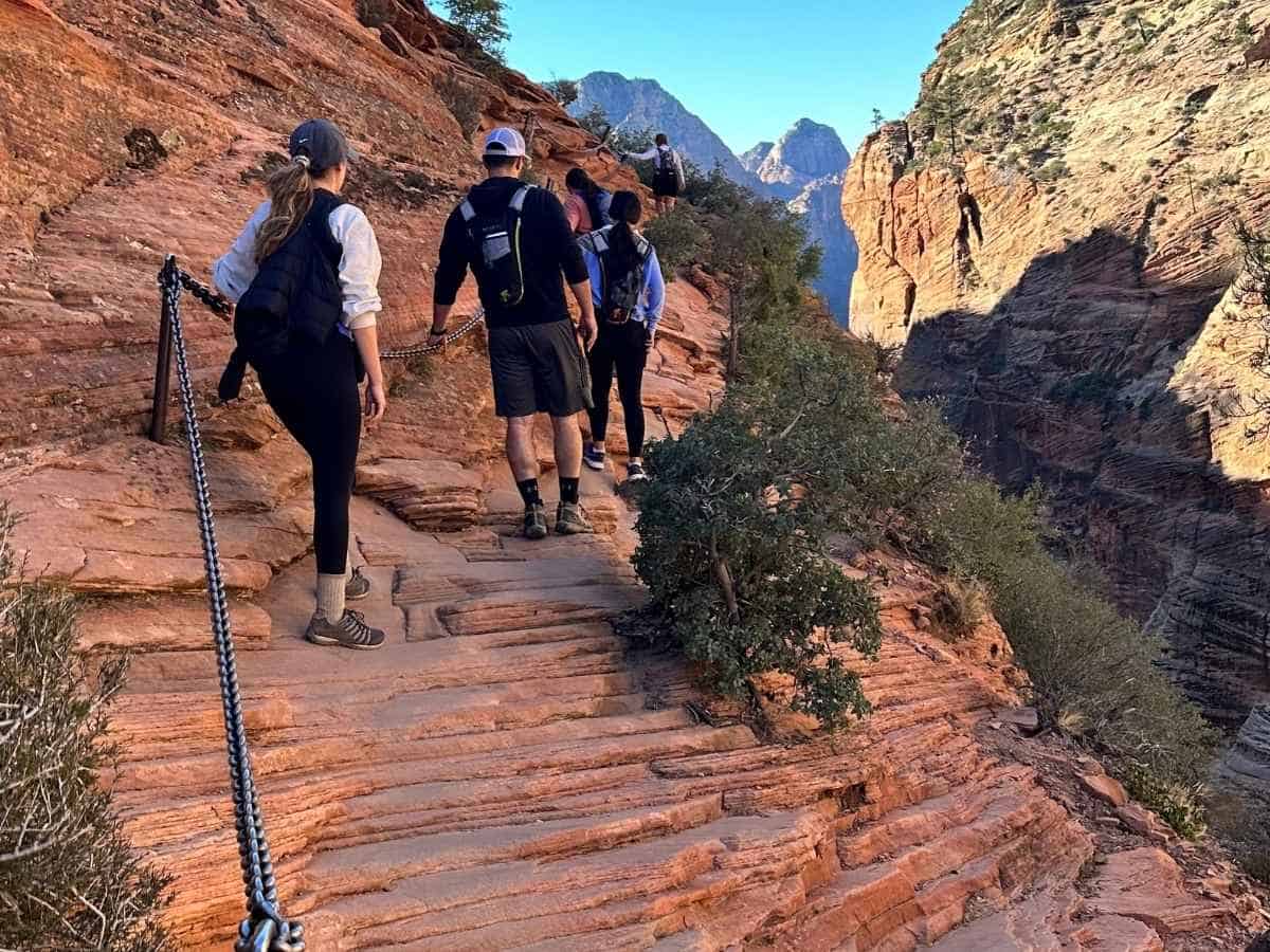 Group of hikers carefully making their way along the narrow Angels Landing trail, gripping the metal safety chain bolted into the red rock cliff. The steep drop offs and carved sandstone path showcase one of the most adventurous highlights to consider in a Zion National Park itinerary.