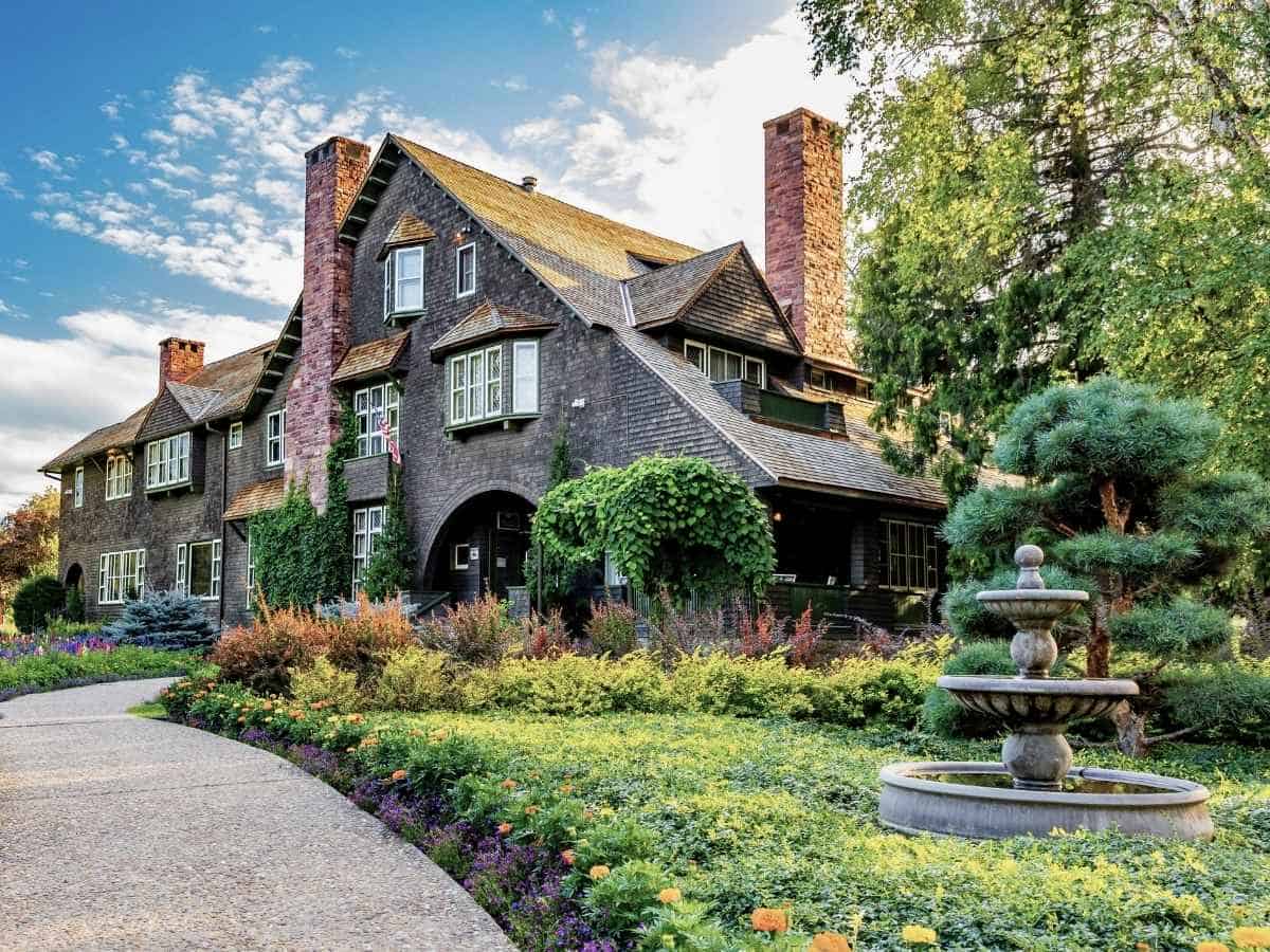 Drive Bozeman to Glacier National Park Road Trip Large historic lodge-style building with dark wood shingles, tall brick chimneys, and ivy climbing the exterior surrounded by colorful gardens and a stone fountain. A charming historic stop travelers may visit while exploring towns along a Bozeman to Glacier National Park road trip.