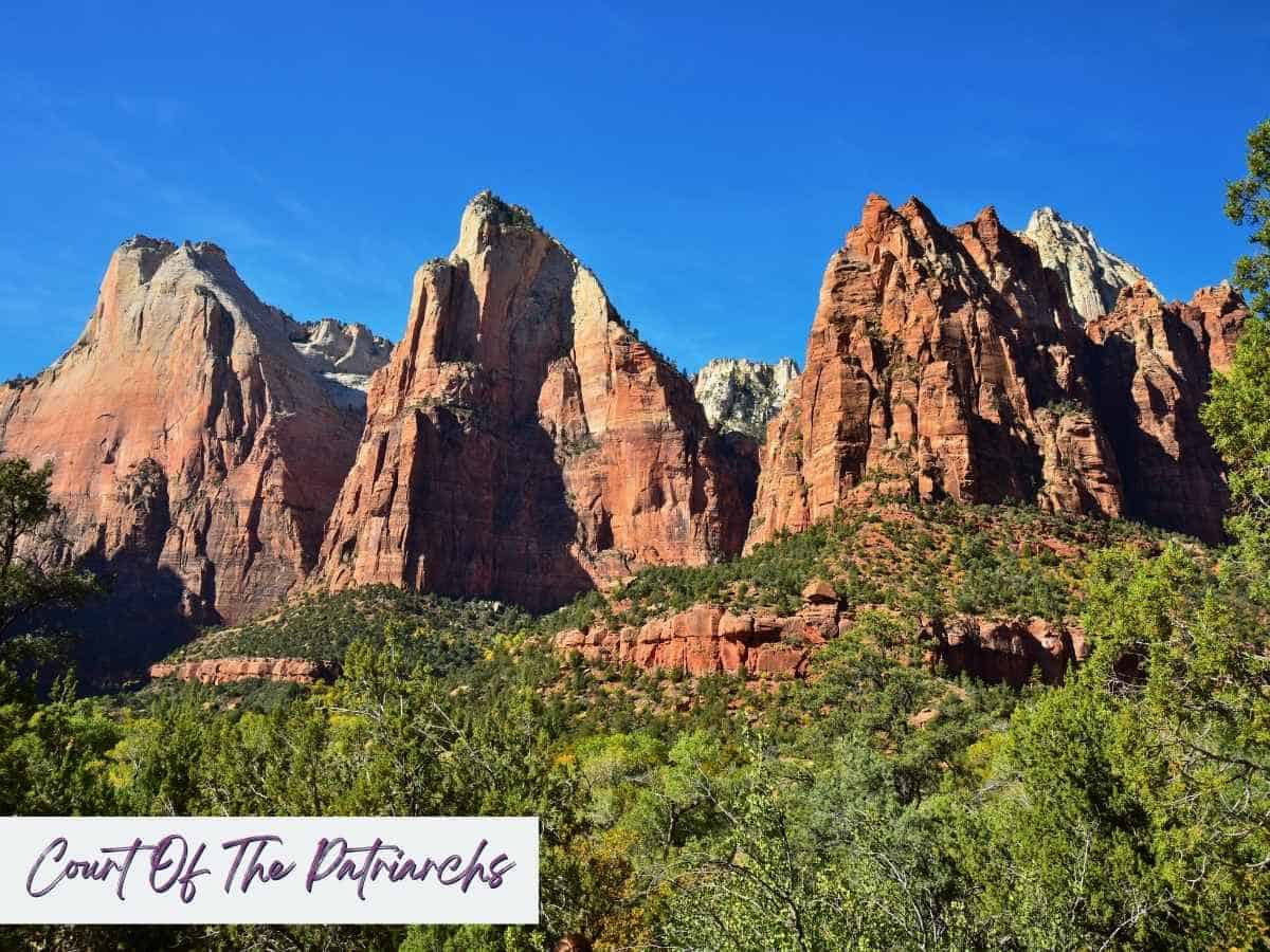 Towering red and cream colored sandstone cliffs rise above lush green trees in the Court of the Patriarchs area of Zion National Park. Text on the image reads Court Of The Patriarchs, highlighting this iconic viewpoint often featured in a Zion National Park itinerary for classic canyon scenery.
