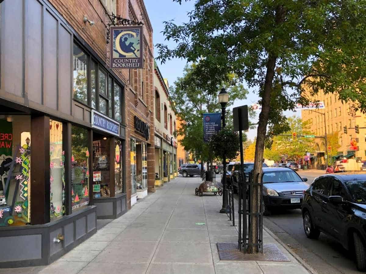 Drive Bozeman to Glacier National Park Road Trip Small town sidewalk lined with local shops including a storefront sign reading "COUNTRY BOOKSHELF" on a brick building with parked cars and leafy trees along the street. A charming downtown stop travelers might explore while passing through Montana on a Bozeman to Glacier National Park road trip.