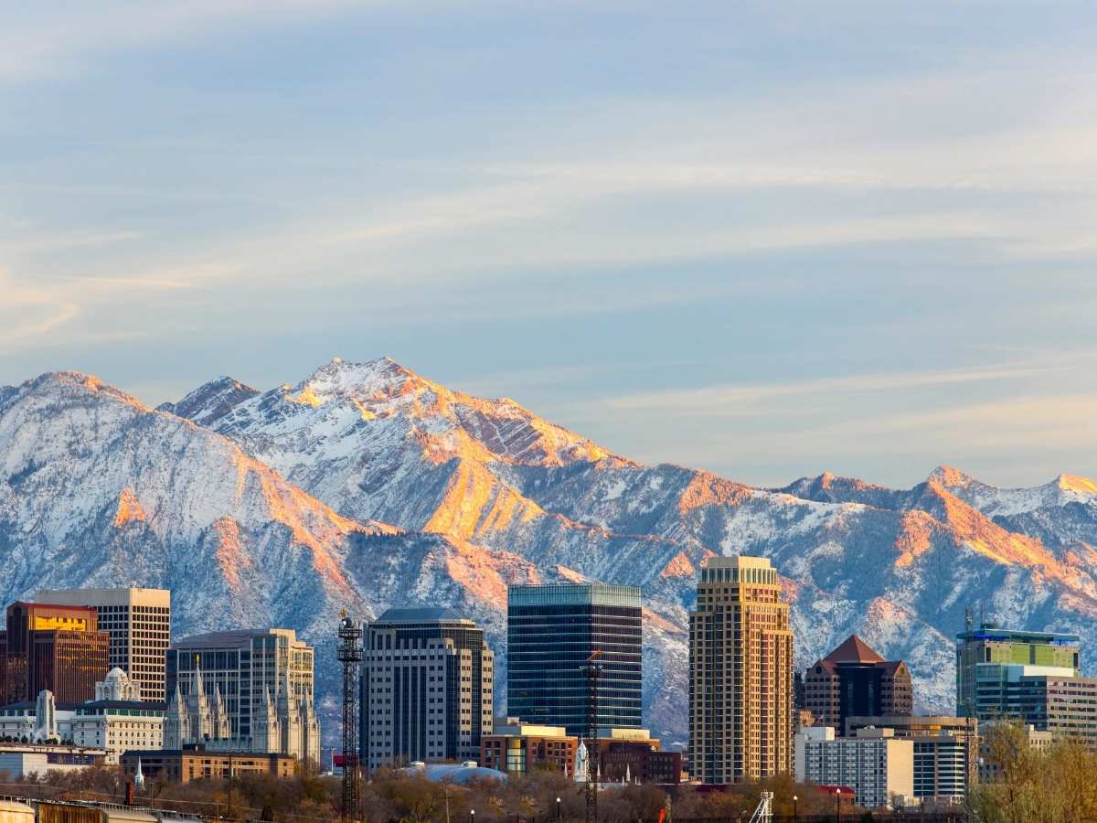 City skyline with modern buildings and a prominent white temple set against snow covered mountains glowing in warm sunset light under a soft sky. Starting point for the drive from Salt Lake City to Arches National Park road trip, showcasing the city framed by the Wasatch Range.