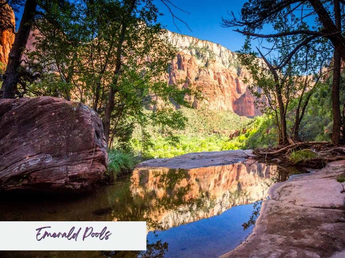 Small reflective pool at Emerald Pools in Zion National Park mirrors the surrounding red rock cliffs and green trees under a bright blue sky. Text on the image reads Emerald Pools, showcasing this peaceful and scenic stop often included in a Zion National Park itinerary for an easy nature walk with beautiful canyon views.