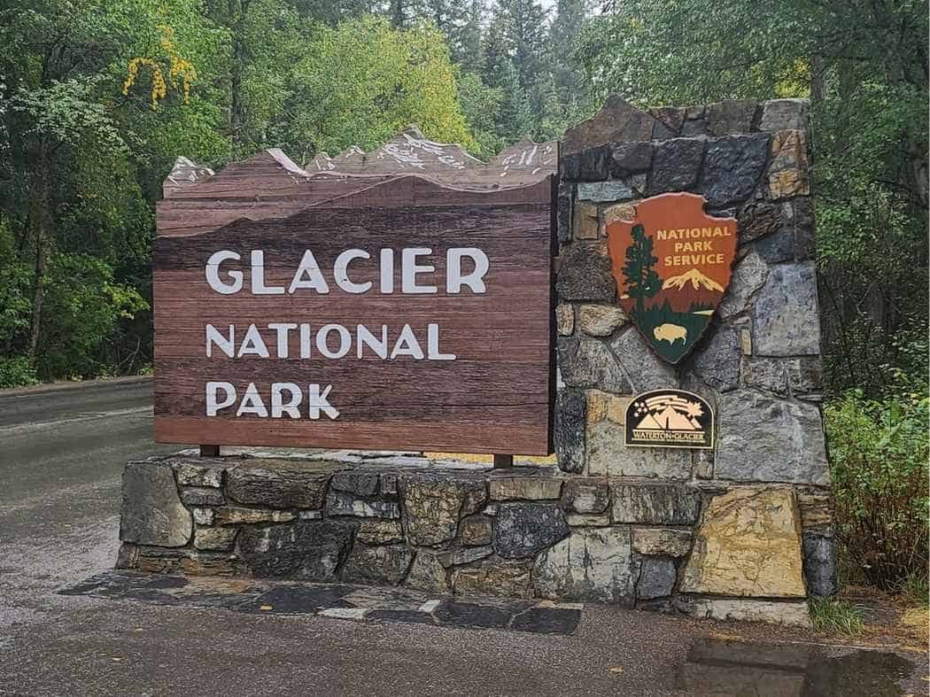Wooden entrance sign beside a stone pillar at the park entrance that reads "GLACIER NATIONAL PARK" with the "NATIONAL PARK SERVICE" emblem and a "WATERTON GLACIER INTERNATIONAL PEACE PARK" plaque. Trees and a roadway surround the sign marking the entrance featured in a Glacier National Park itinerary.