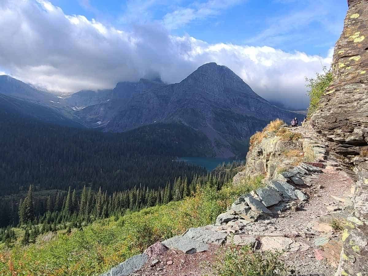 Rocky hiking trail carved into a cliff in Many Glacier with a sweeping view of forested valleys and rugged mountains in Glacier National Park. Low clouds drift around the peaks while hikers stand on the narrow path overlooking the dramatic landscape featured in a Glacier National Park itinerary.