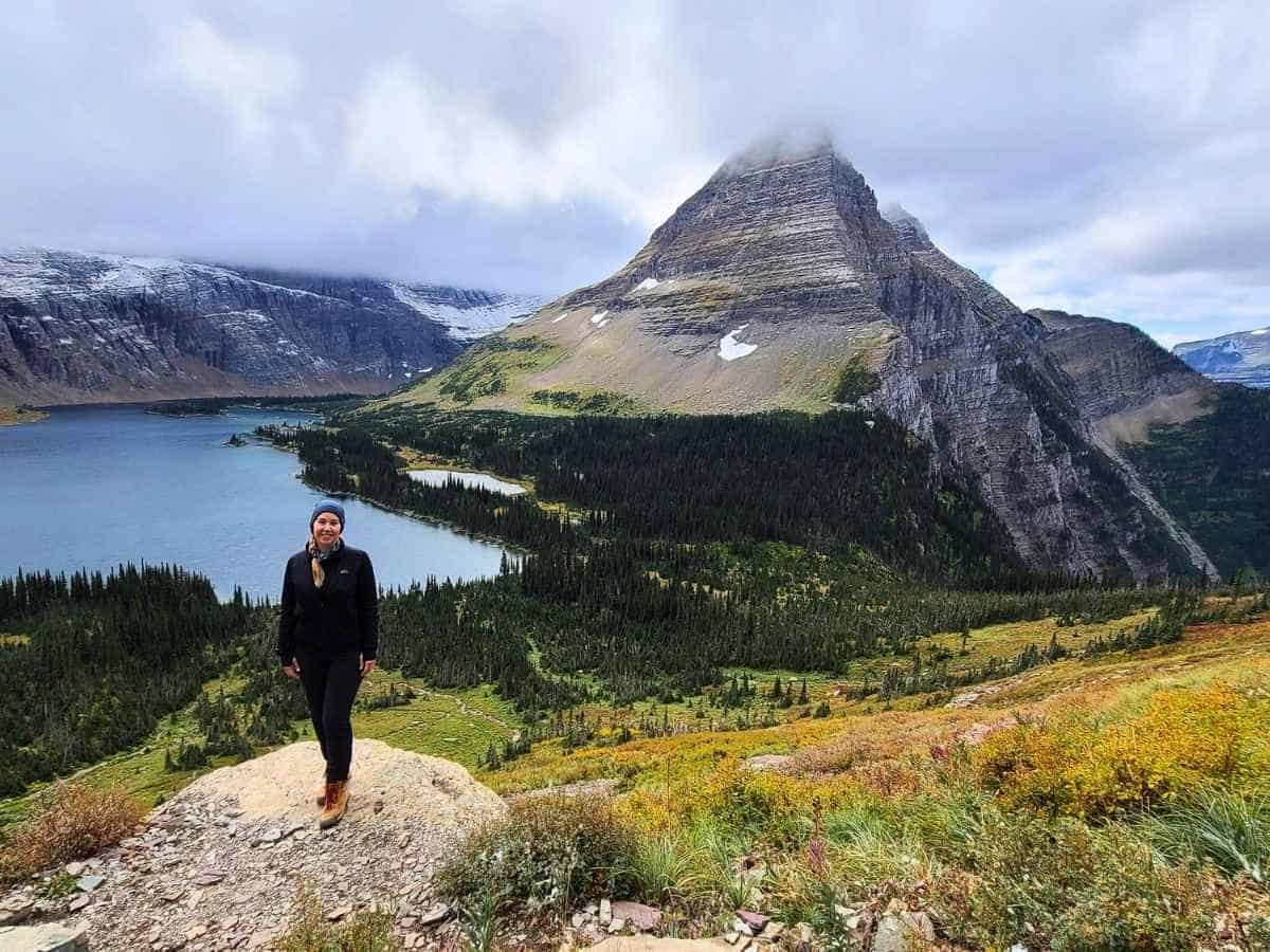 Woman standing on a rocky viewpoint overlooking a long alpine lake surrounded by dense evergreen forest and steep mountains in Glacier National Park. Low clouds hang over the peaks while the scenic overlook highlights a stop along Going-to-the-Sun Road in a Glacier National Park itinerary.
