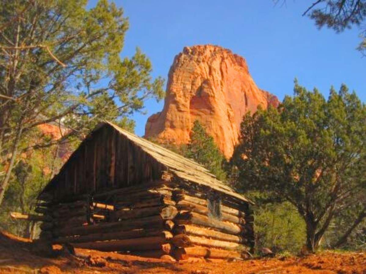 Rustic log cabin sits among red dirt and green trees with a towering sandstone cliff glowing in warm sunlight behind it in Zion National Park. This charming historic style cabin adds a cozy and scenic lodging option to consider in a Zion National Park itinerary.