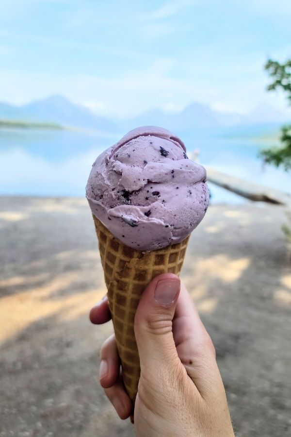 Hand holding a waffle cone with a scoop of purple huckleberry ice cream in the foreground with a calm lake and hazy mountain peaks in the background. A sweet roadside treat enjoyed during a scenic stop on a Bozeman to Glacier National Park road trip.