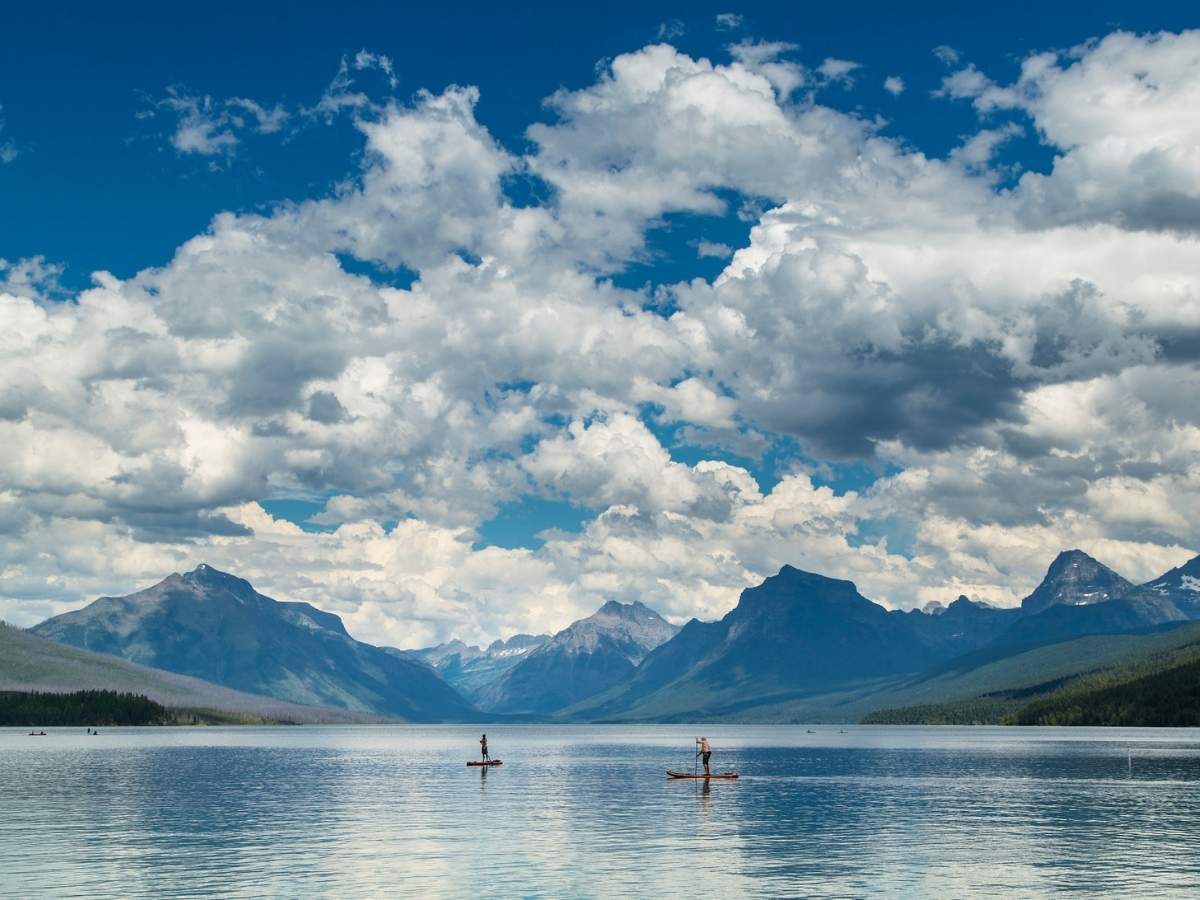 Wide view of a calm mountain lake with two people paddleboarding on the water surrounded by rugged peaks and dramatic clouds in Glacier National Park. The expansive lake and mountain scenery highlight outdoor activities included in a Glacier National Park itinerary.