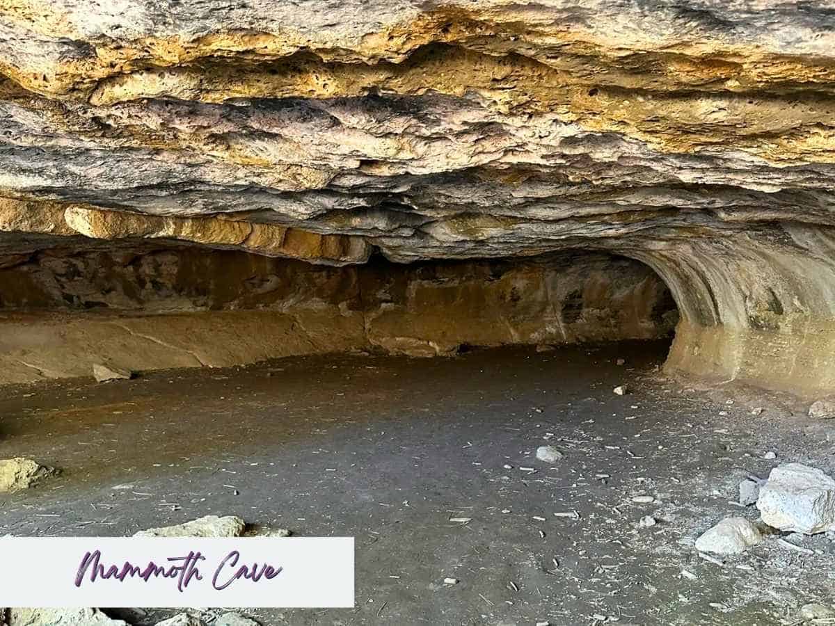 Low wide limestone cave interior with layered rock ceiling and a curved tunnel like opening carved into the wall at Mammoth Cave in Utah. The natural cave formation highlights an underground stop that can be explored along the route from Zion to Bryce Canyon National Park with the text “Mammoth Cave” written in the bottom corner.