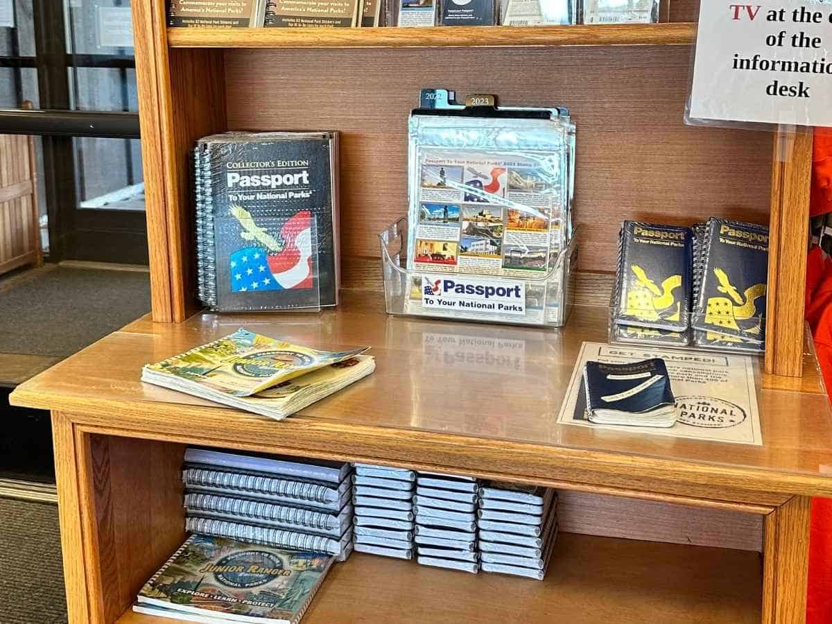 Display shelf filled with booklets and souvenirs including multiple copies of "Passport to Your National Parks" and "Junior Ranger" books arranged in stacks. Visitor center stop along the drive from Salt Lake City to Arches National Park road trip, highlighting where to pick up a national parks passport book and stamps.