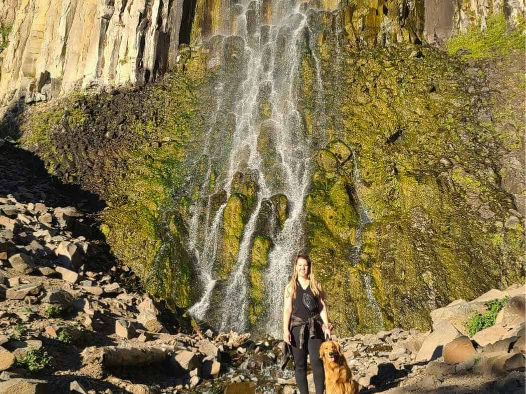 Drive Bozeman to Glacier National Park Road Trip Tall waterfall streaming down a steep rocky cliff covered in green moss with a woman standing at the base beside a golden retriever on the rocky ground. A scenic waterfall stop that highlights the dramatic landscapes travelers can discover on a Bozeman to Glacier National Park road trip.