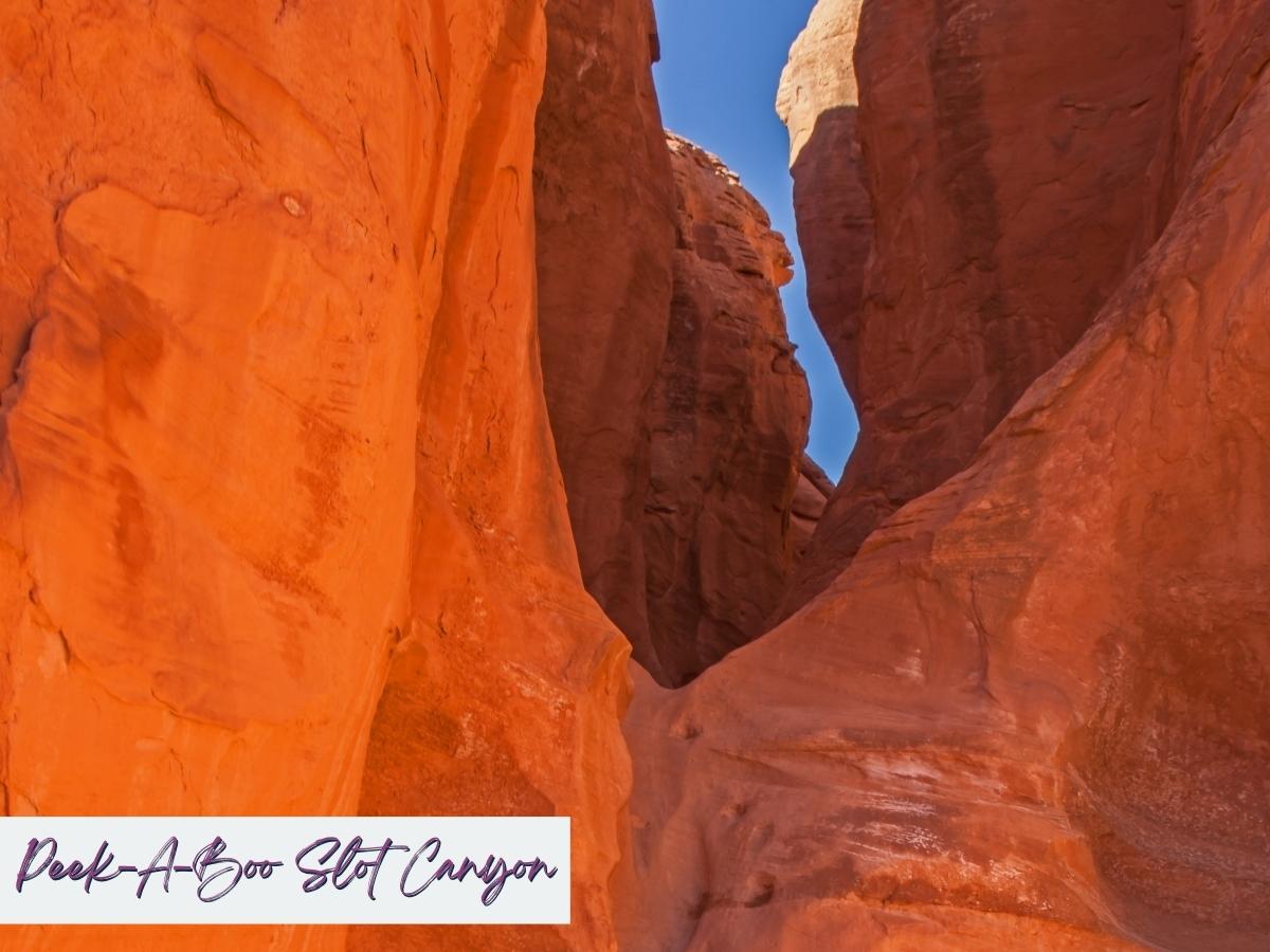 Tall narrow red sandstone walls rise closely together inside Peek A Boo Slot Canyon with sunlight illuminating the smooth curved rock surfaces and a strip of blue sky above. The winding canyon passage highlights a dramatic slot canyon stop often explored on trips from Zion to Bryce Canyon National Park with the text “Peek A Boo Slot Canyon” written in the bottom corner.