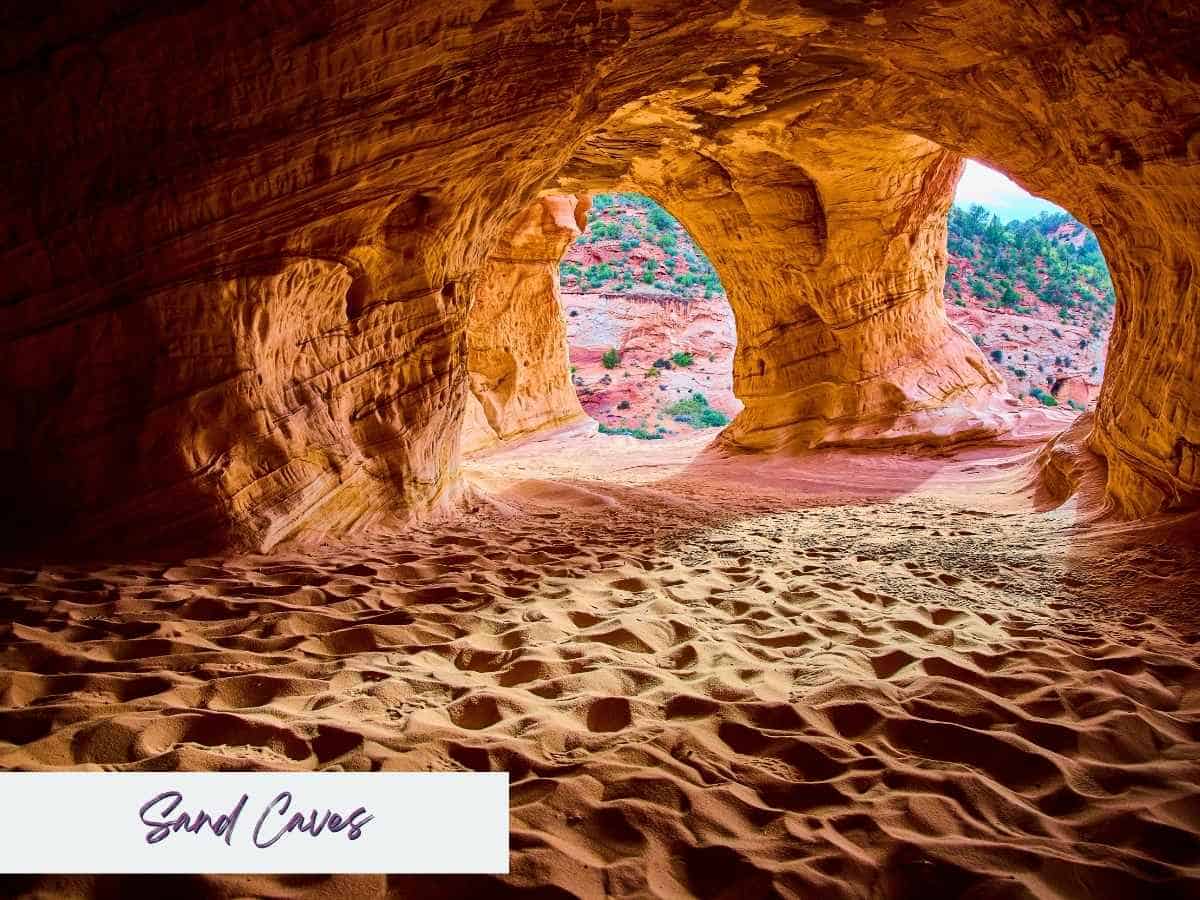 View from inside large red sandstone sand caves near Kanab Utah with rippled sand covering the ground and two wide cave openings looking out to desert cliffs and scattered green trees. The unique cave formation highlights a scenic stop often visited on the drive from Zion to Bryce Canyon National Park with the text “Sand Caves” written in the bottom corner.