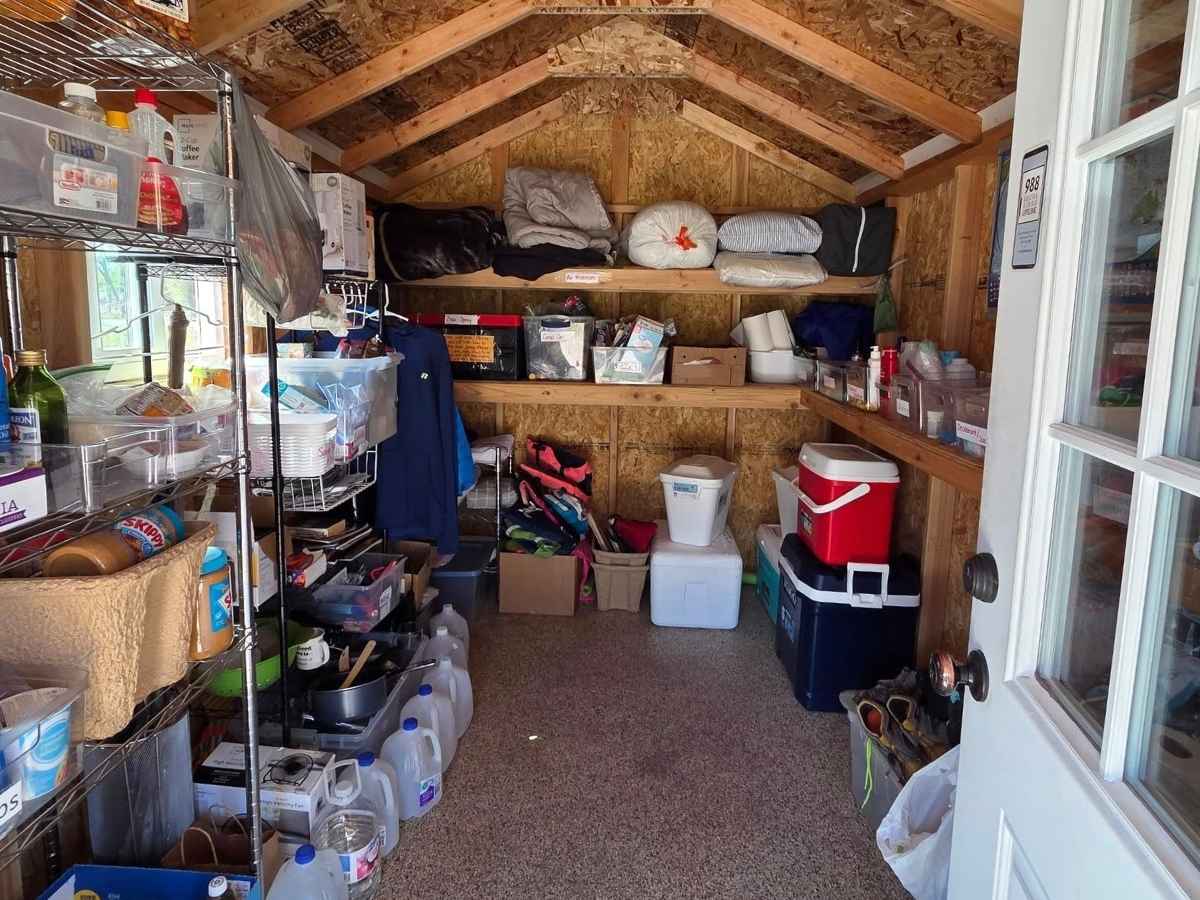 Small wooden shed organized with camping and road trip supplies including coolers, water jugs, cookware, blankets, and food containers stacked on shelves. A practical packing and supply setup for preparing for a Bozeman to Glacier National Park road trip.