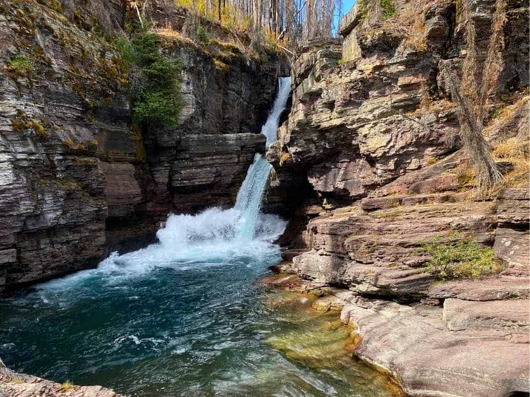 Waterfall cascading through a narrow rocky canyon into a turquoise pool surrounded by layered stone cliffs in Glacier National Park. The scenic waterfall stop highlights St Mary or Virginia Falls along Going-to-the-Sun Road in a Glacier National Park itinerary.