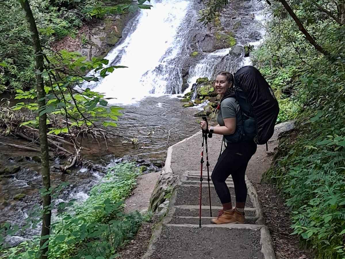 Drive Bozeman to Glacier National Park Road Trip Hiker with a large backpack and trekking poles standing on stone steps beside a rushing waterfall surrounded by dense green forest. Trekking poles are an essential packing item for a Bozeman to Glacier National Park road trip.