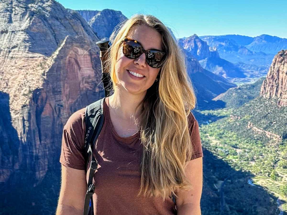 Smiling hiker wearing sunglasses and a backpack stands at a scenic overlook high above Zion Canyon, with towering red rock cliffs and the winding valley below. This close up viewpoint shot captures the breathtaking scale and unforgettable scenery that make a Zion National Park itinerary so memorable.