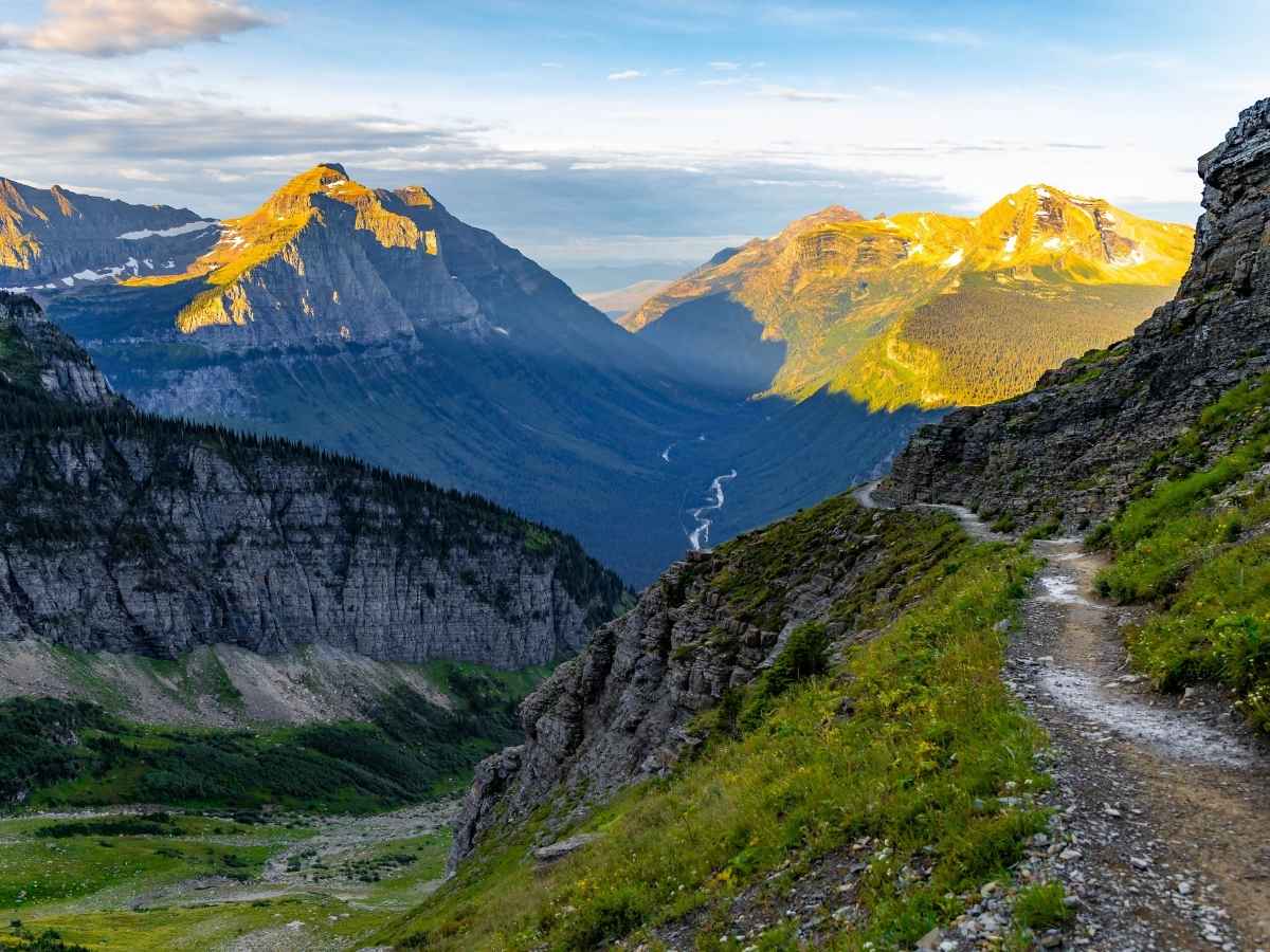 Narrow hiking trail along a steep mountainside in Glacier National Park overlooking a wide valley with a winding river and sunlit peaks in the distance. This scenic viewpoint on Highline trail highlights the rugged terrain visitors can access after using glacier national park shuttle tickets to reach trailheads.