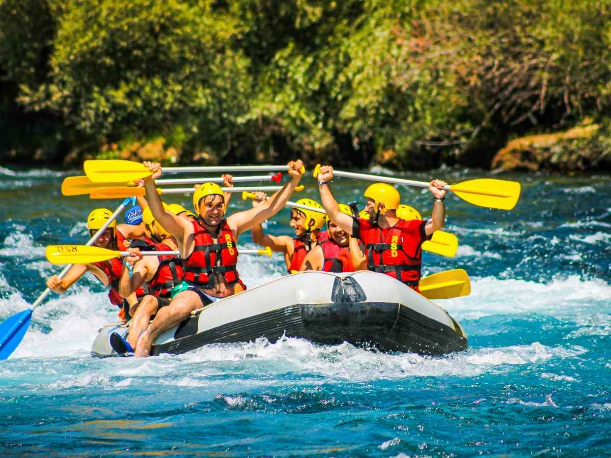 Drive Bozeman to Glacier National Park Road Trip Group of people wearing yellow helmets and red life jackets paddling an inflatable raft through bright blue river rapids with paddles raised in celebration. An adventurous whitewater rafting stop that can add excitement to a Bozeman to Glacier National Park road trip.