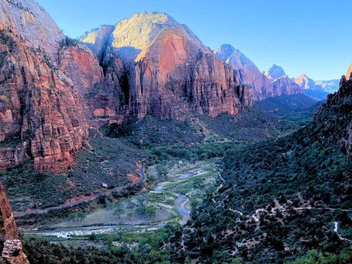 Wide view of Zion National Park canyon with towering red rock cliffs glowing in soft light above a winding river and road through the green valley below. This scenic overlook captures the dramatic landscape and scale that make a Zion National Park itinerary unforgettable for first time visitors.