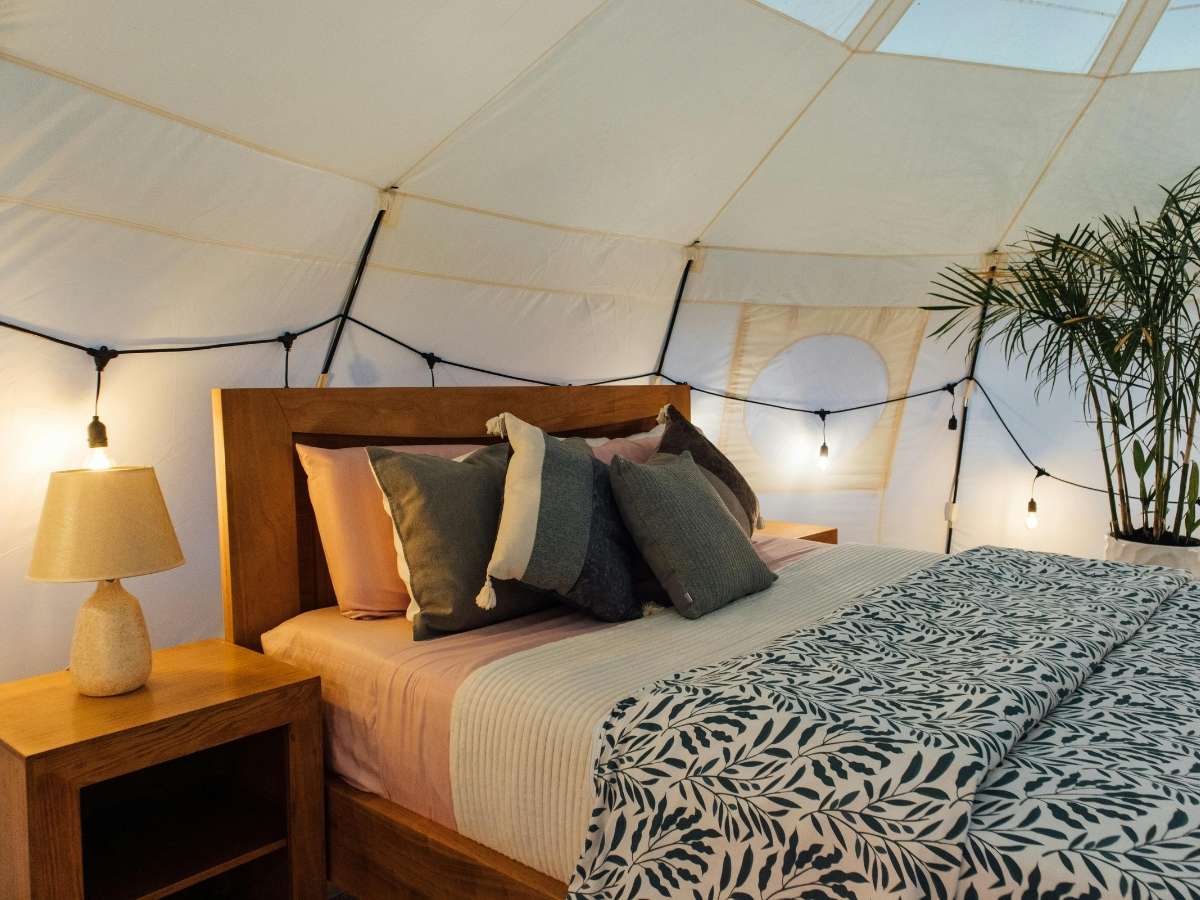 Cozy glamping tent interior with a wooden bed frame, soft neutral bedding, and layered throw pillows under warm string lights. This stylish and comfortable stay offers a unique lodging option to include in a Zion National Park itinerary for a memorable desert experience.