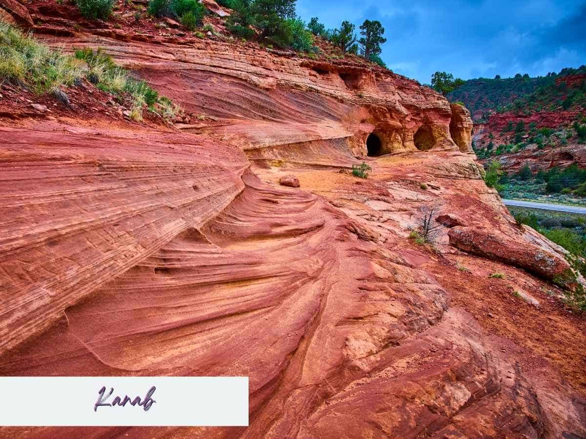 Layered red sandstone cliffs with smooth wave like formations and small cave openings carved into the rock along the roadside near Kanab. The striking desert landscape highlights a scenic stop often seen on the drive from Zion to Bryce Canyon National Park with the word “Kanab” written in the bottom corner.