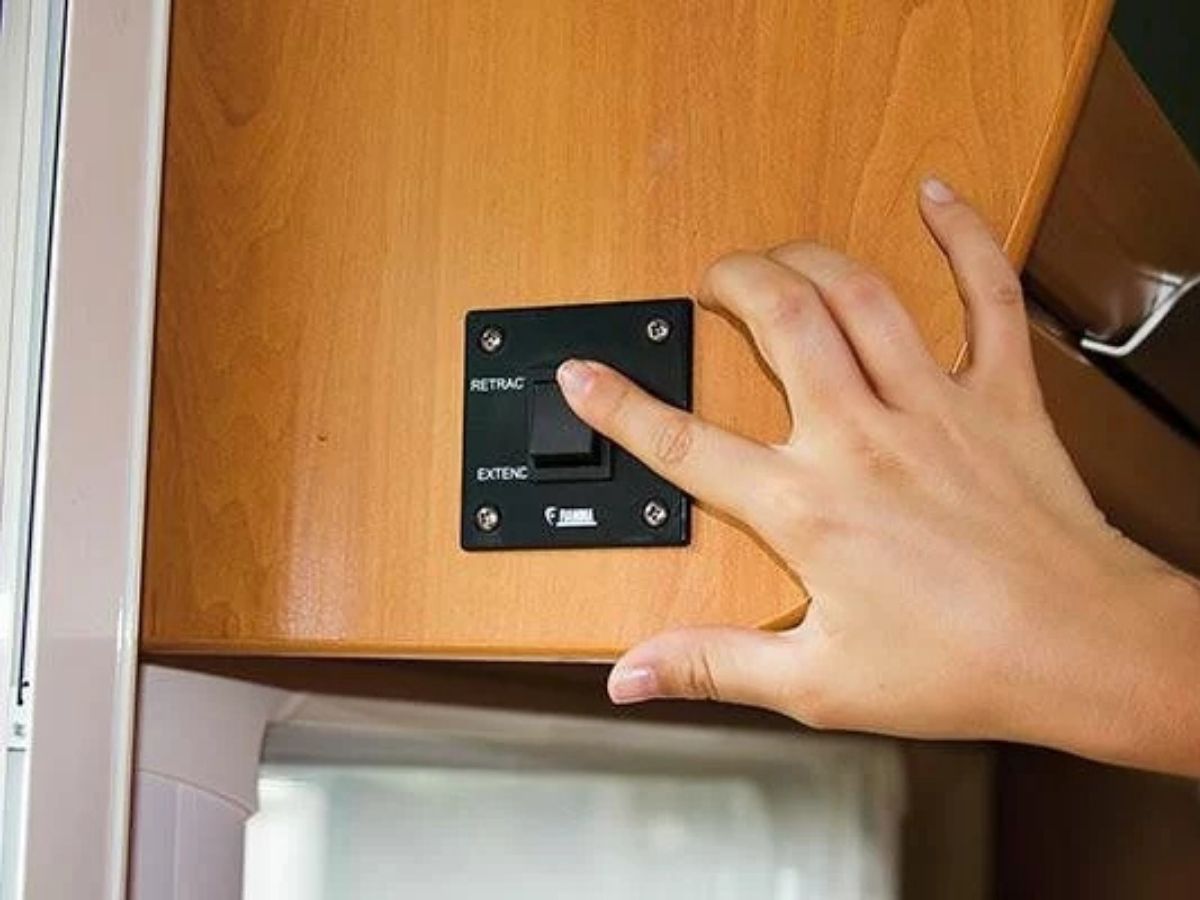 Hand pressing a wall mounted control switch inside a camper van cabinet to operate an awning system. Close up shows labeled buttons reading "RETRACT" and "EXTEND" with "FIAMMA" branding, relevant to using a Fiamma F45S vs F80S awning control.