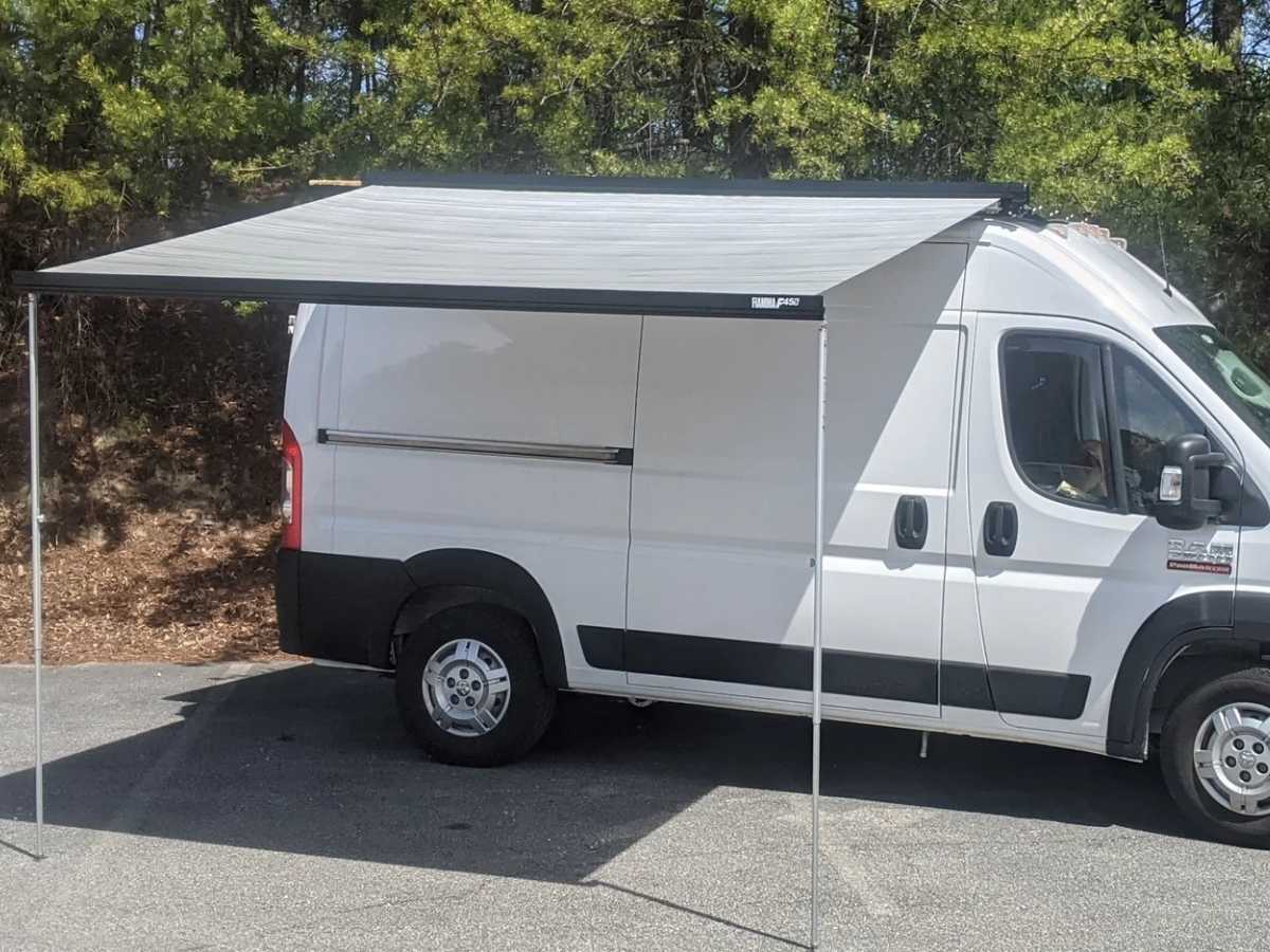 Best Camper Van Awning White camper van parked on a paved area with a fully extended Fiamma F45S camper van awning creating a wide shaded space along the side. Two support poles hold the awning in place while trees line the background. The setup shows a simple outdoor shelter solution for rest stops or camping.
