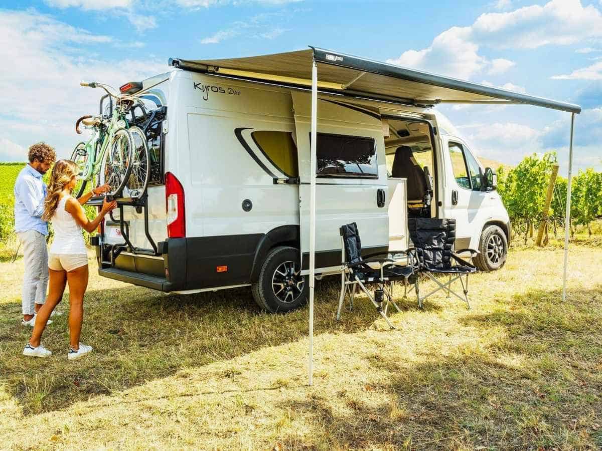 Camper van parked in a sunny field with an extended F80S awning providing shade over two folding chairs and a small outdoor setup. A couple stands at the rear loading bicycles onto a rack, showing a road trip scene that highlights how a Fiamma F45S vs F80S awning enhances outdoor living space.