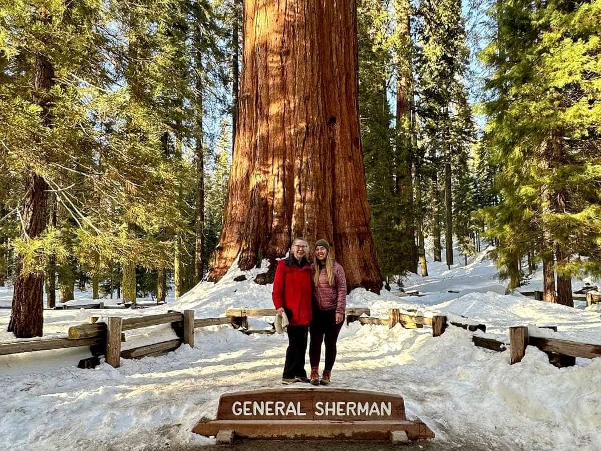 Sequoia National Park Itinerary And Kings Canyon Two visitors stand beside the "General Sherman" sign at the base of the General Sherman Tree during winter, surrounded by snow-covered ground and towering sequoias. This iconic stop is a highlight of a Sequoia National Park itinerary, showcasing the world's largest tree by volume in the heart of Giant Forest.